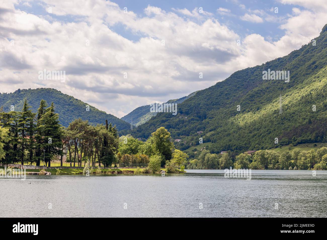 Sonnenlicht erhellt bewaldete Hänge über dem Idrosee in Lombardei, Italien, mit Nadelhölzern und Laubbäumen entlang des Wassers unter einem bewölkten Frühlingshimmel. Stockfoto