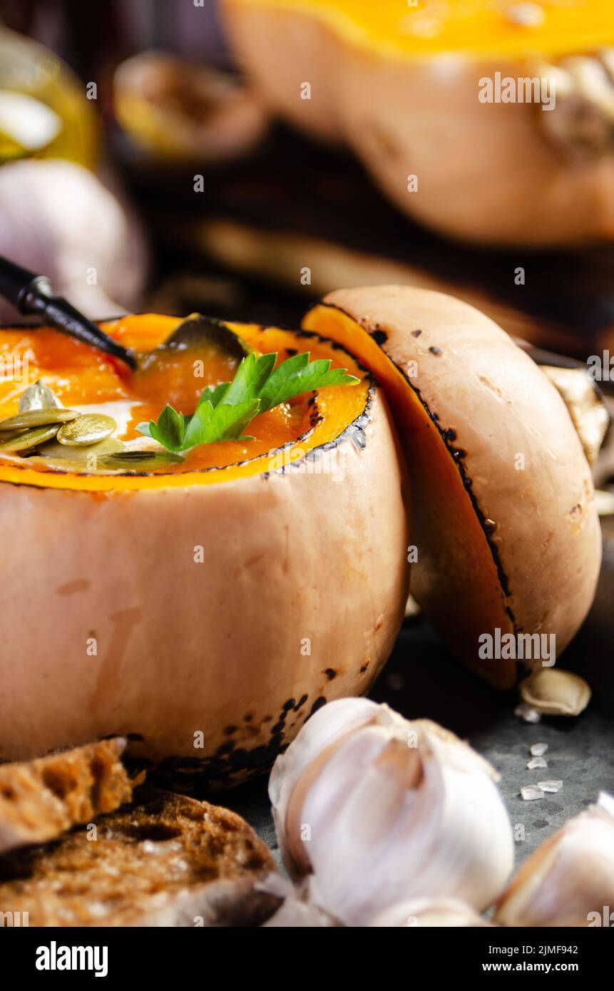 Kürbisschale mit hausgemachter rustikaler Kürbissuppe mit Samen auf Metallschale mit Knoblauch und Brot zur Seite Stockfoto