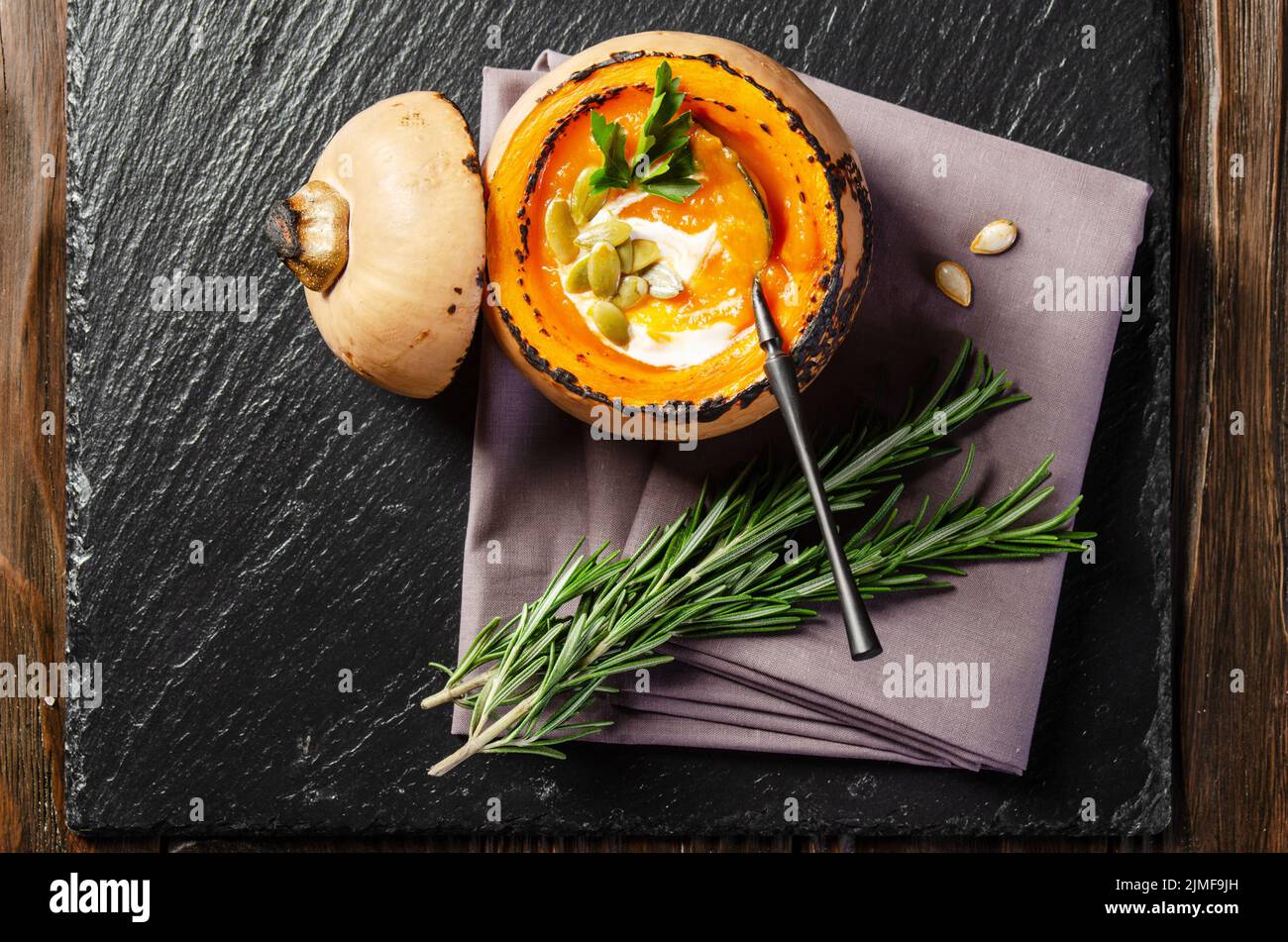 Flacher Laienblick in der Pumpkin-Schale mit hausgemachter rustikaler Kürbissuppe mit Samen auf Schiefertablett mit Rosmarinbeiseite Stockfoto