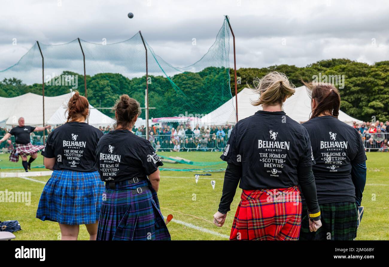 North Berwick, East Lothian, Schottland, Großbritannien, 6.. August 2022. North Berwick Highland Games: Die jährlichen Spiele finden auf dem Erholungsgebiet statt. Im Bild: Weibliche Teilnehmer der Schwerevents tragen Kilts und T-Shirts, die von der Belhaven Brewery gesponsert werden, und sehen einer Teilnehmerin beim Shot Put-Event zu Stockfoto