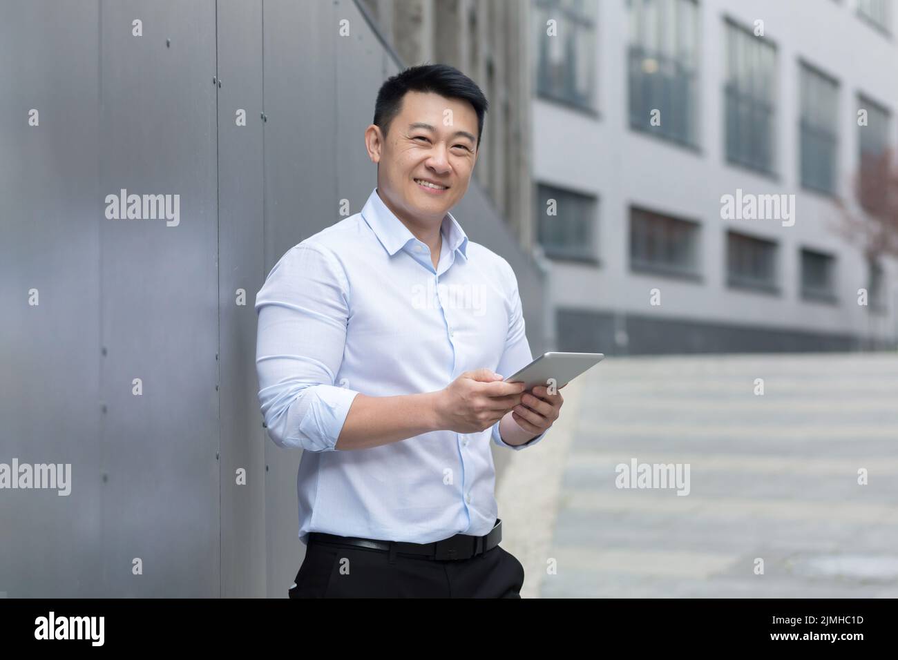 Erfolgreicher asiatischer Geschäftsmann mit Tablet-Computer in den Händen, lächelnd und mit Blick auf die Kamera, Mann, der vor dem Bürogebäude steht Stockfoto