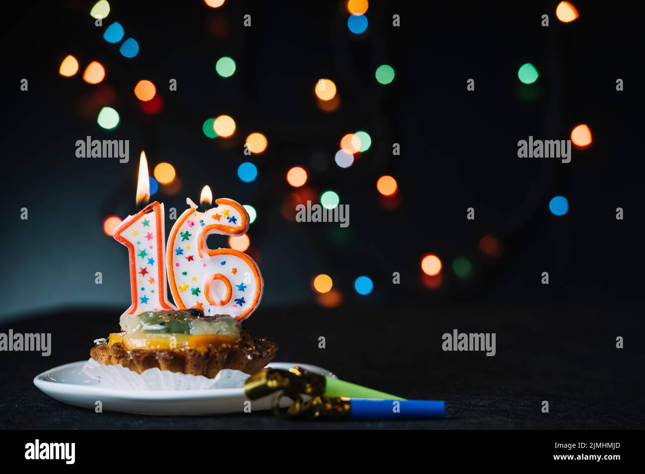 Die Zahl 16 Geburtstag beleuchtete Kerze in Scheiben mit Party-Horn-Gebläse vor beleuchtetem Bokeh-Hintergrund Stockfoto