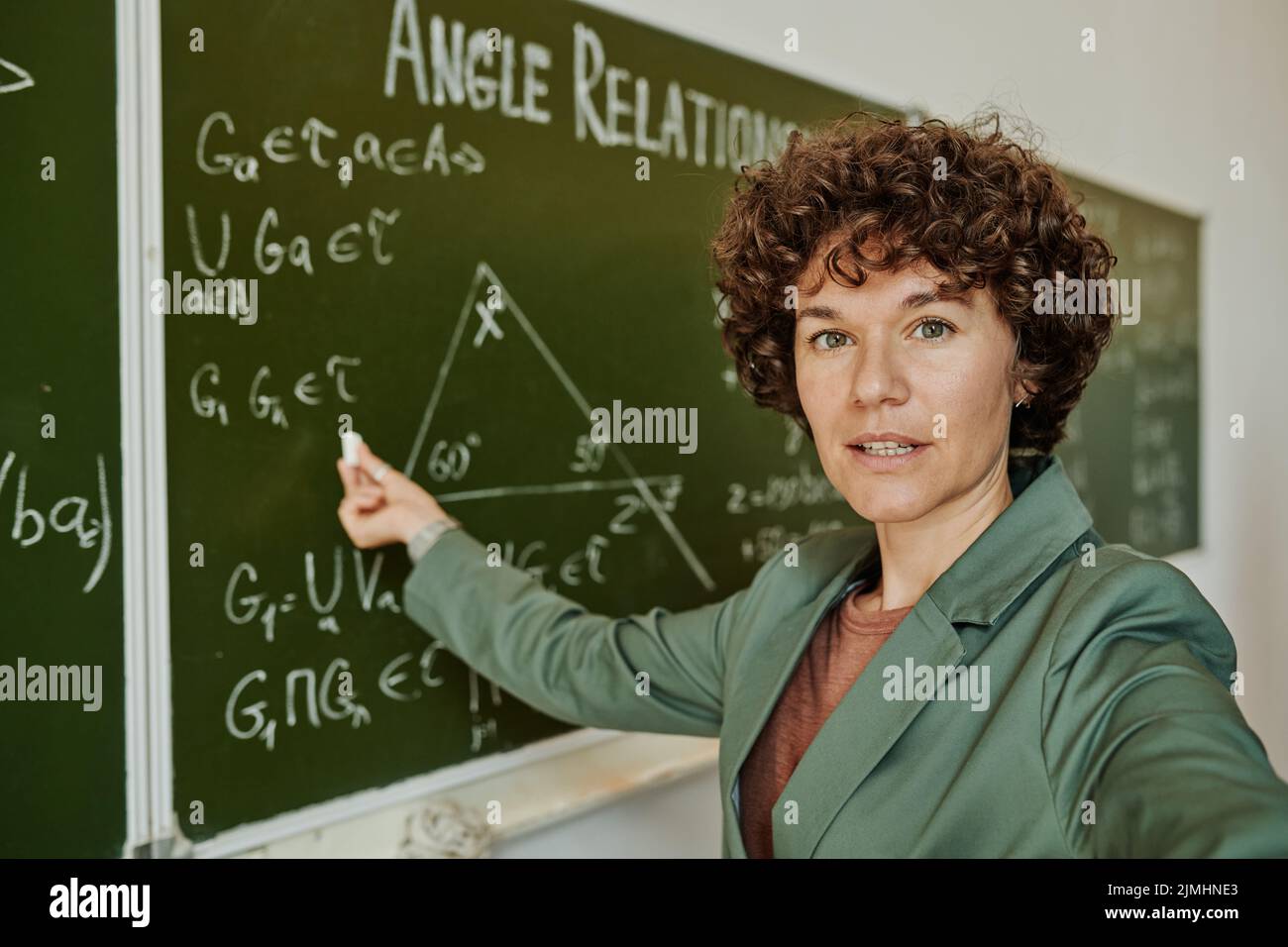 Selbstbewusster Geometrielehrer in formalwear zeigt auf Tafel mit Formeln und Dreieck während der Erklärung für Online-Publikum Stockfoto