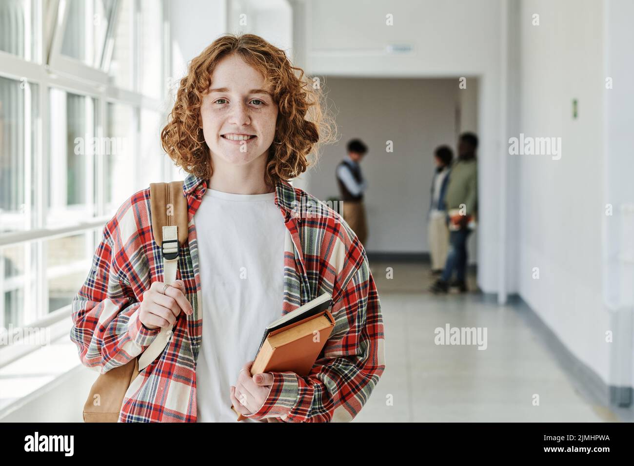 Nette, erfolgreiche Teenager-Studentin mit Büchern und Rucksack, die auf die Kamera schauen, während sie im Flur gegen eine Gruppe von Klassenkameraden steht Stockfoto