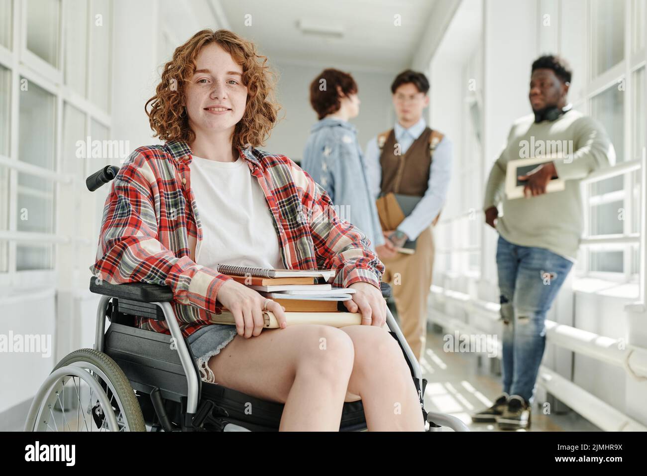 Lächelndes Teenager-Mädchen mit körperlicher Behinderung in Casualwear, das im Rollstuhl im College-Korridor gegen eine Gruppe von Klassenkameraden sitzt Stockfoto