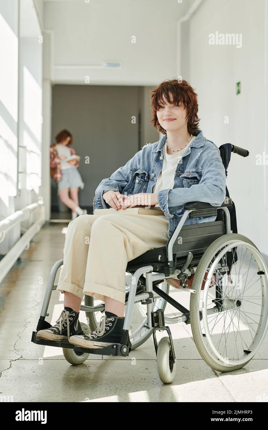 Glückliche Teenager-Studentin mit körperlicher Behinderung, die im Rollstuhl im College-Korridor sitzt und die Kamera gegen ihren Klassenkameraden anschaut Stockfoto