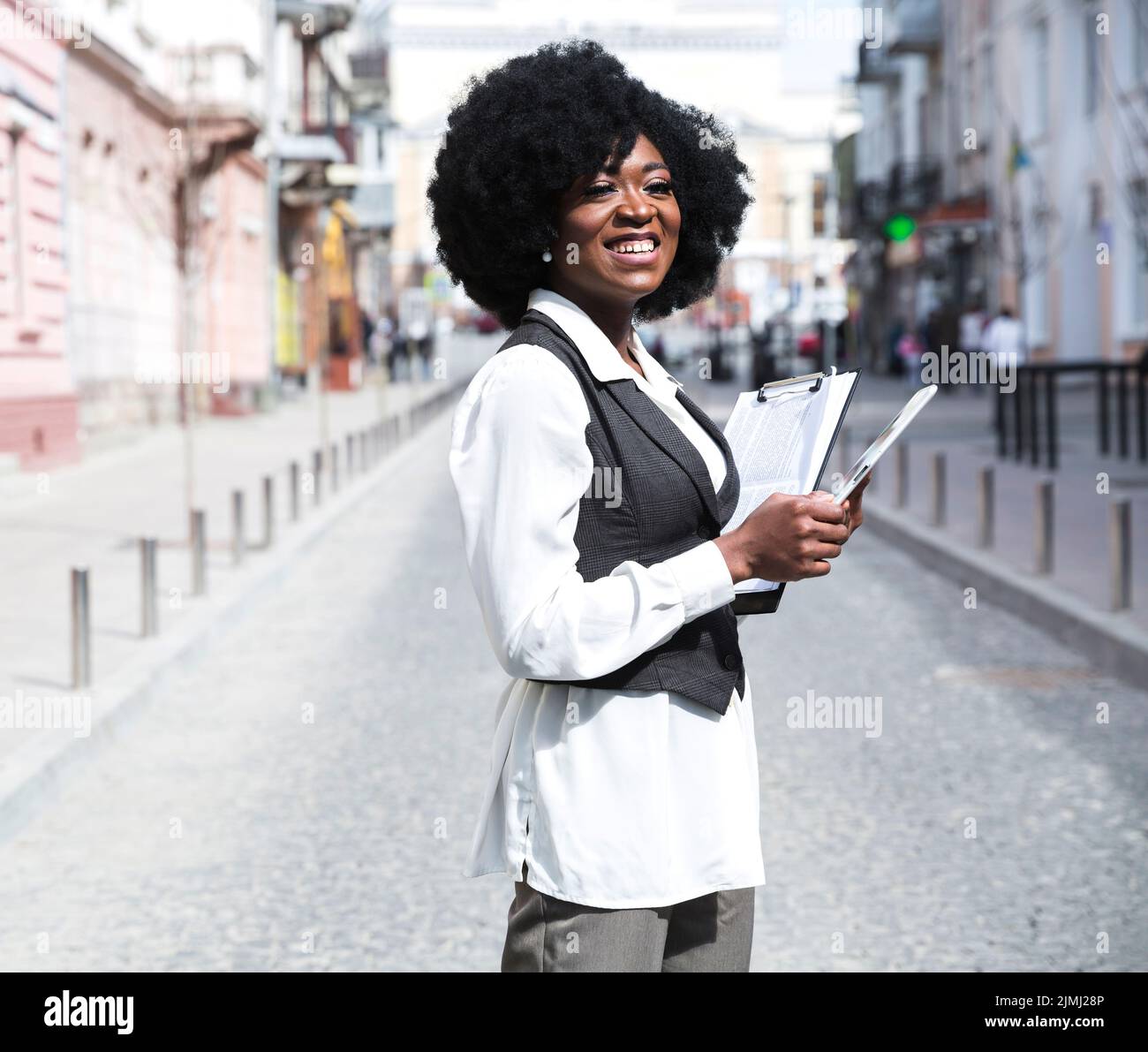 Lächelnde junge afrikanische Geschäftsfrau steht Stadt Straße hält Zwischenablage wegschauen Stockfoto