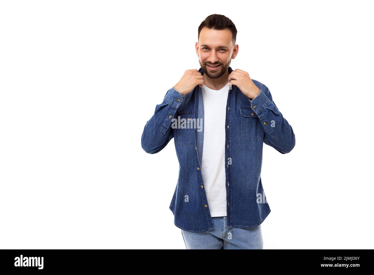Stilvoller, eleganter Mann in einem weißen T-Shirt und einem hellblauen Denim-Hemd richtet die Kleidung am Kragen aus und blickt lächelnd auf die Kamera Stockfoto