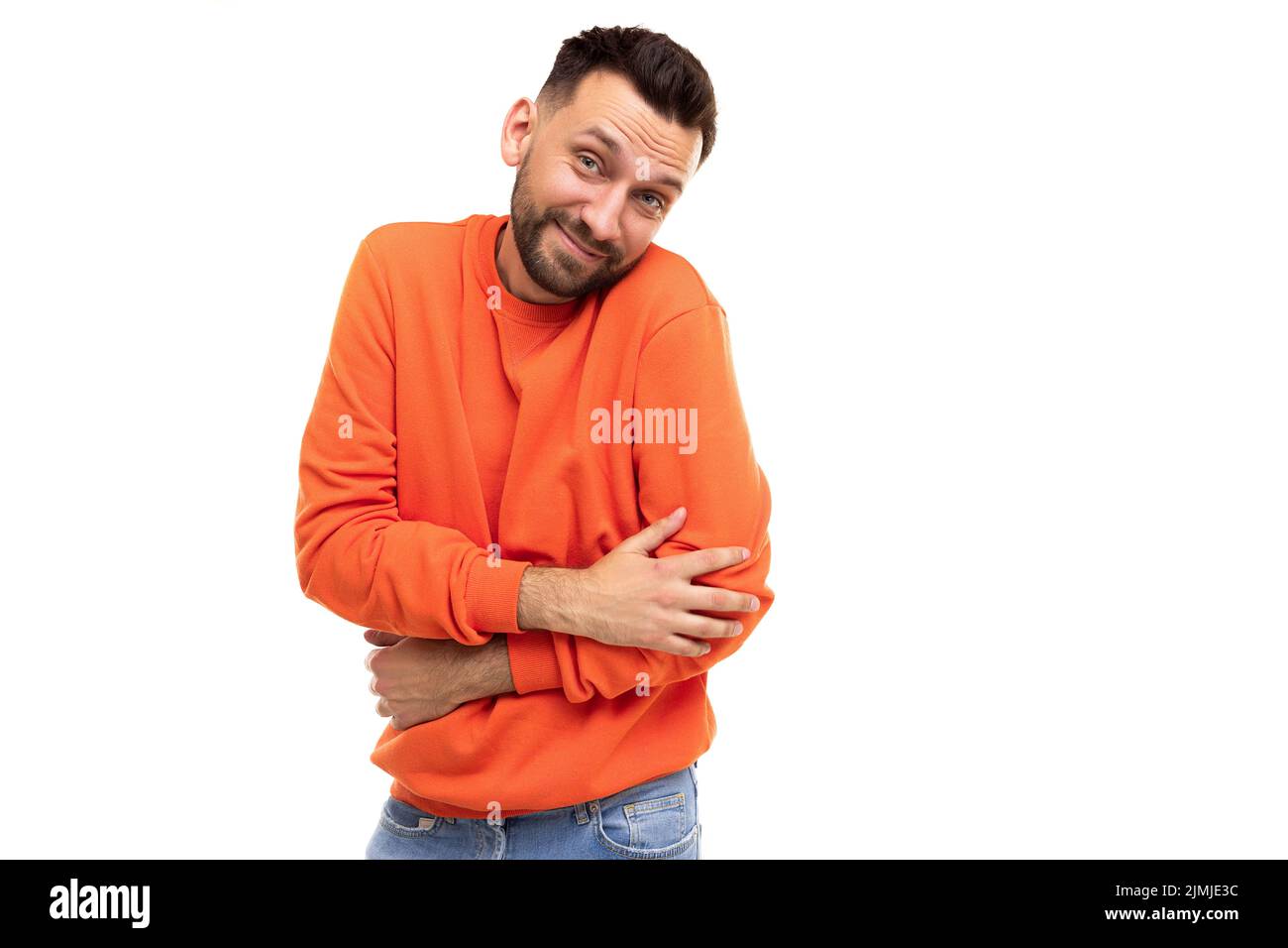 Schüchterner bärtiger Mann in einem stilvollen orangefarbenen Langarm schließt schüchtern seine Hände vor dem Hintergrund einer weißen Wand Stockfoto