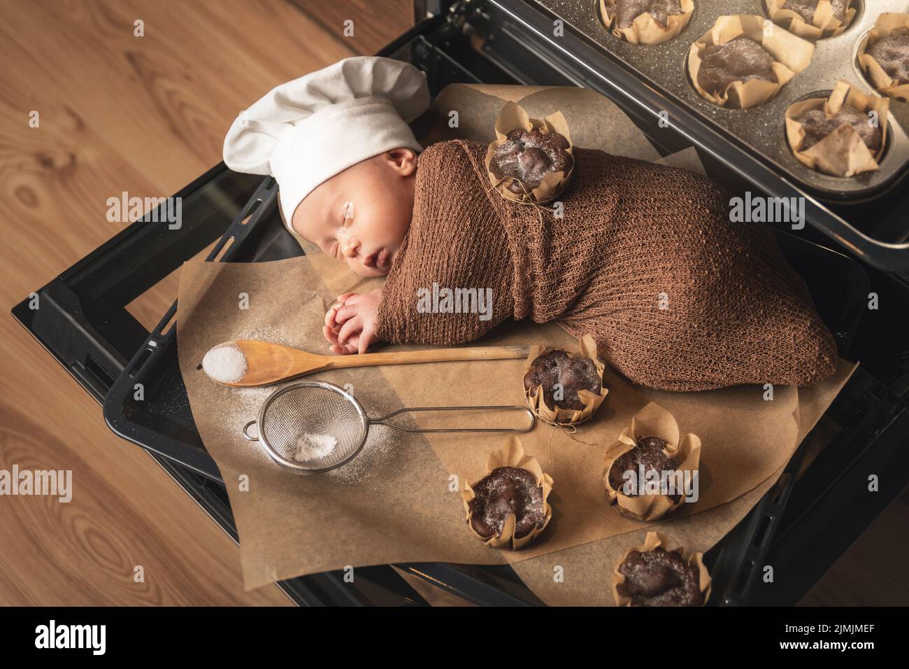 Das neugeborene Baby mit Kochmütze liegt mit Muffins auf dem Backblech Stockfoto