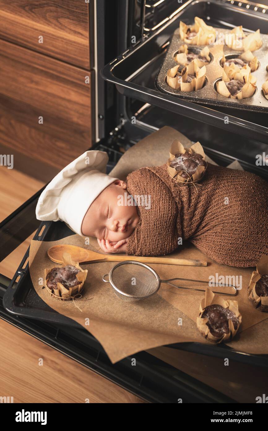 Das neugeborene Baby mit Kochmütze liegt mit Muffins auf dem Backblech Stockfoto
