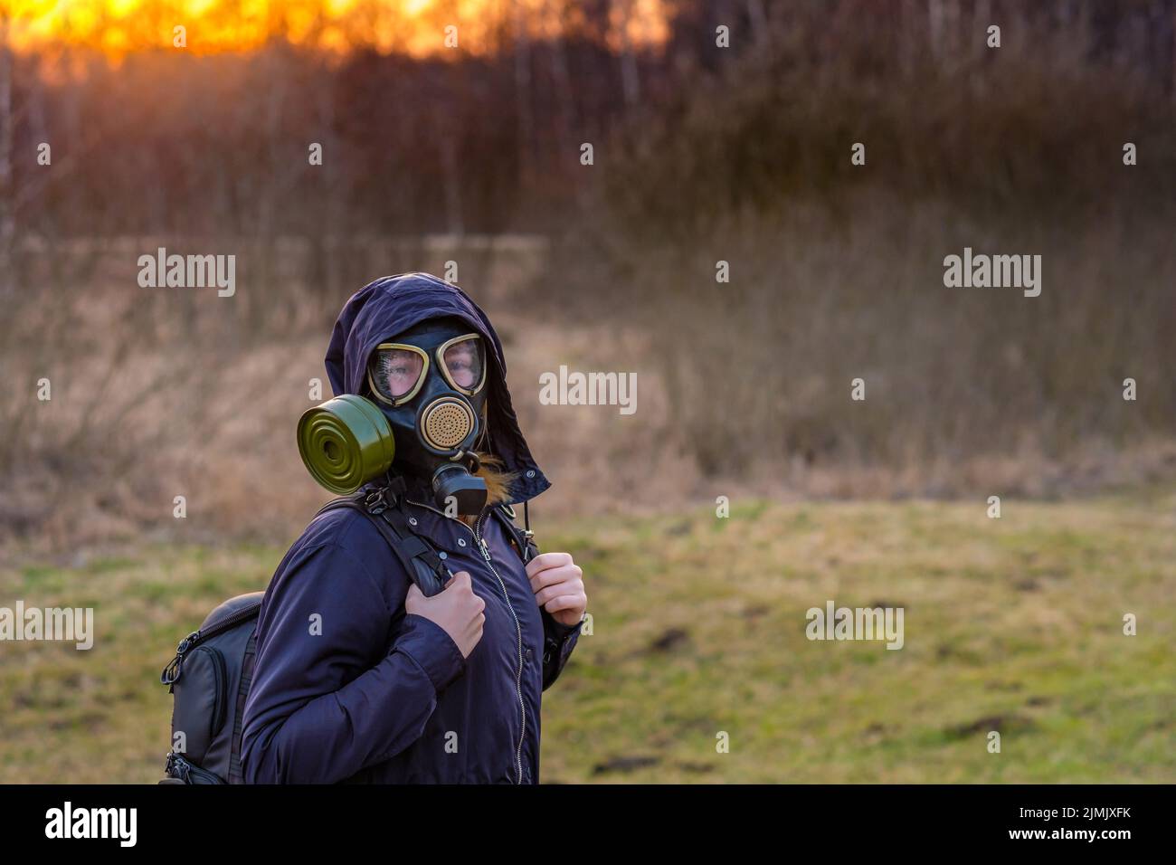 Ein Mädchen in einer schwarzen Gasmaske und dunkler Kleidung mit einer Kapuze bei Sonnenuntergang vor dem Hintergrund eines Frühlingswaldes. Stockfoto