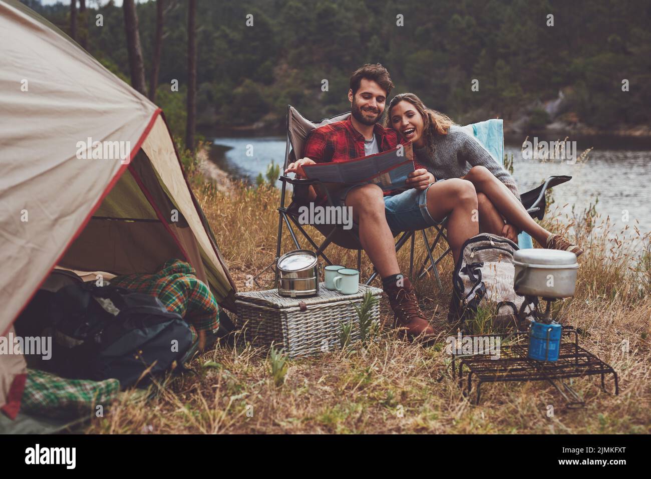 Liebevolles junges Paar, das eine Reisekarte liest, während es vor dem Zelt sitzt. Glückliches junges Paar, das neben dem See zeltet. Stockfoto