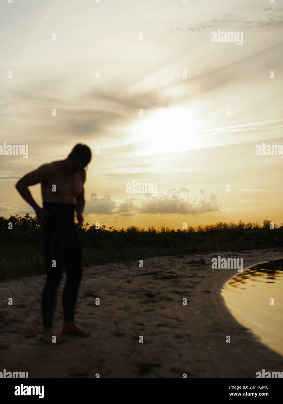 Taucherin, der einen Neoprenanzug aufsetzt, während er bei Sonnenuntergang am Strand steht. Abenteuerlicher junger Mann, der sich darauf vorbereitet, am Meer ins Wasser zu kommen. Stockfoto