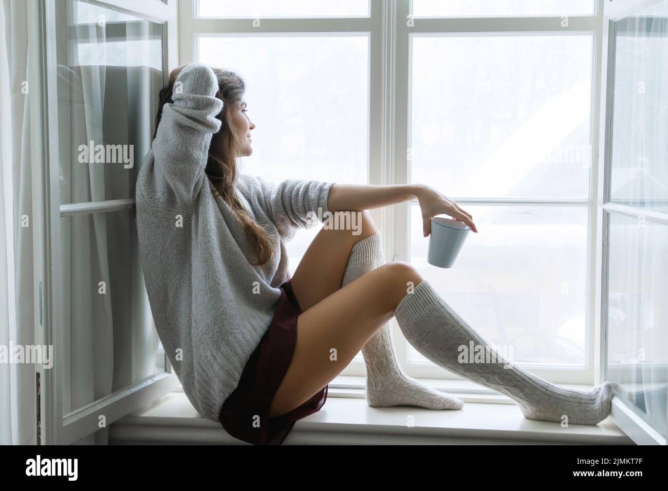 Frau sitzt auf der Fensterbank und trinkt heißen Tee oder Kaffee Stockfoto