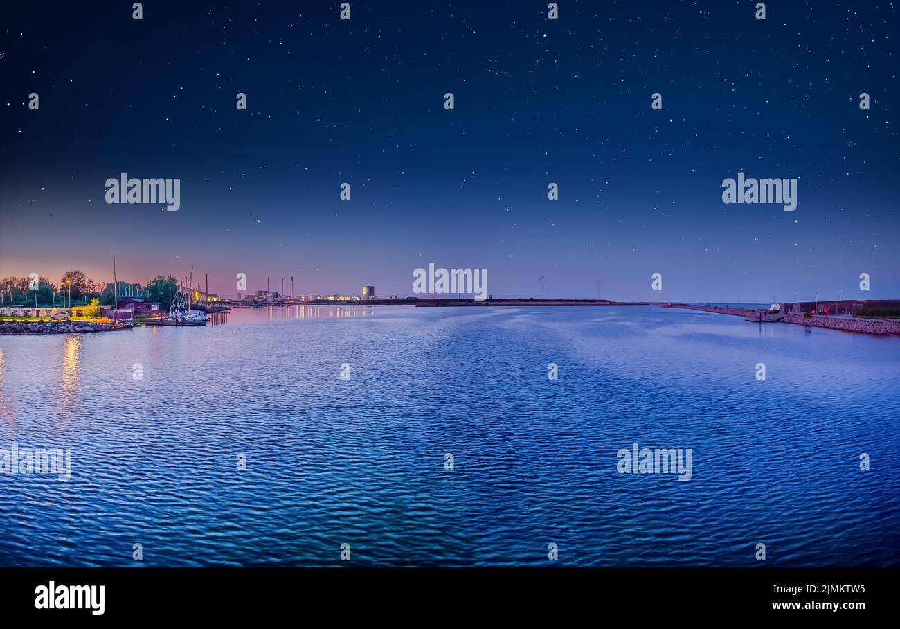 Eine Bucht mit blauem Wasser, eine Graslandinsel PrÃ¸vestenen Syd mit kleinen Häusern, Windturbinen und einer Fabrik vor dem Hintergrund Stockfoto