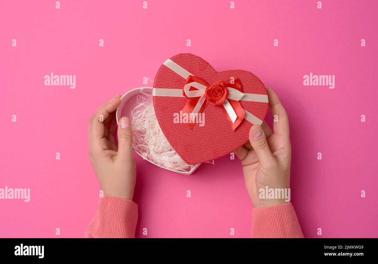 Zwei weibliche Hände halten eine rote herzförmige Box auf einem rosa Hintergrund. Festlicher Hintergrund Stockfoto