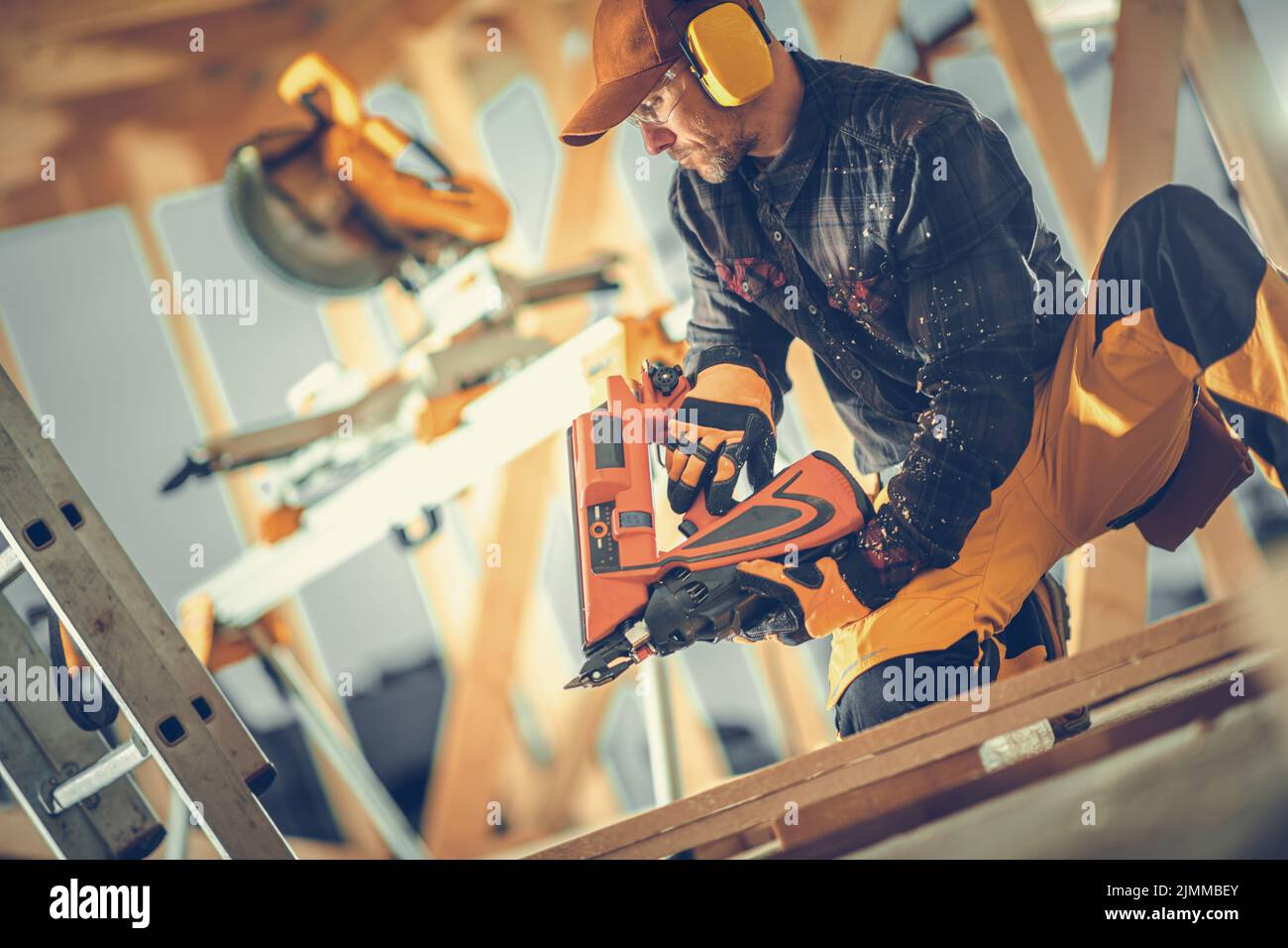 Kaukasischer Bauarbeiter in Sicherheitskopfhörern Bau eines Holzrahmens eines Wohnhauses im kanadischen Stil mit Nail Gun Tool. Industriedesign. Stockfoto