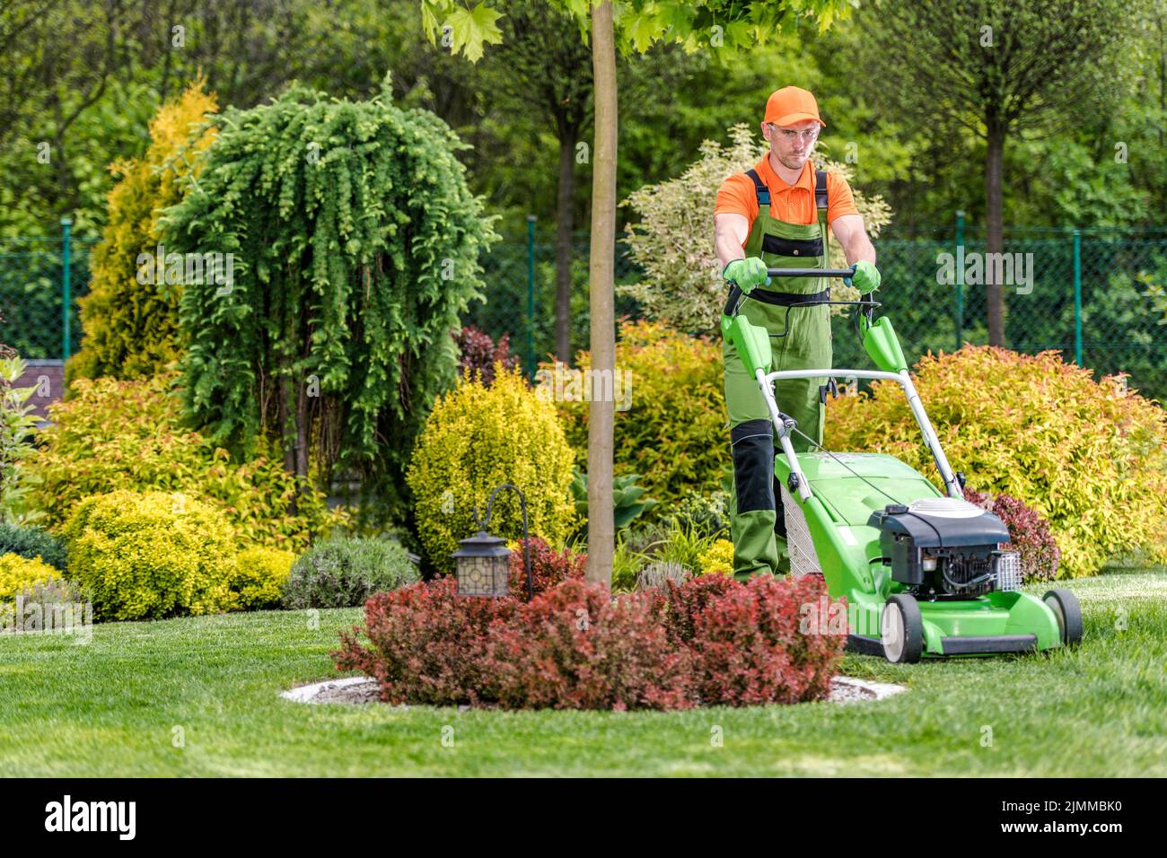 Kaukasischer Greenskeeper trimmt das Gras im Garten seines Kunden mit elektrischem Rasenmäher. Wunderschön angelegte Pflanzen im Hintergrund. G Stockfoto