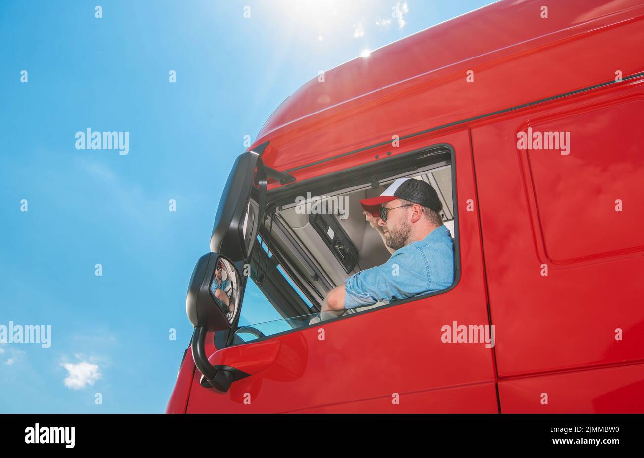 LKW-Fahrer in einem blauen Hemd und Baseballmütze, der in seinem riesigen Reg-Lastwagen sitzt und die Straße vor sich sieht. Blauer sonniger Himmel im Hintergrund. Transportation Stockfoto