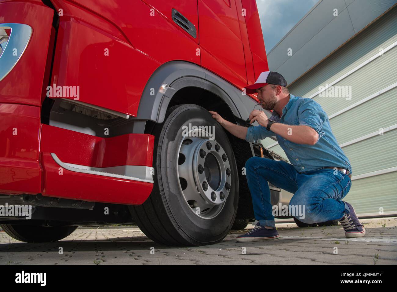 Caucasian Trucker überprüft den Zustand der Reifen in seinem großen Heavy Duty Red Semi Truck, bevor er sich zur Lieferung einer Ladung aufsetzt. Transporta Stockfoto