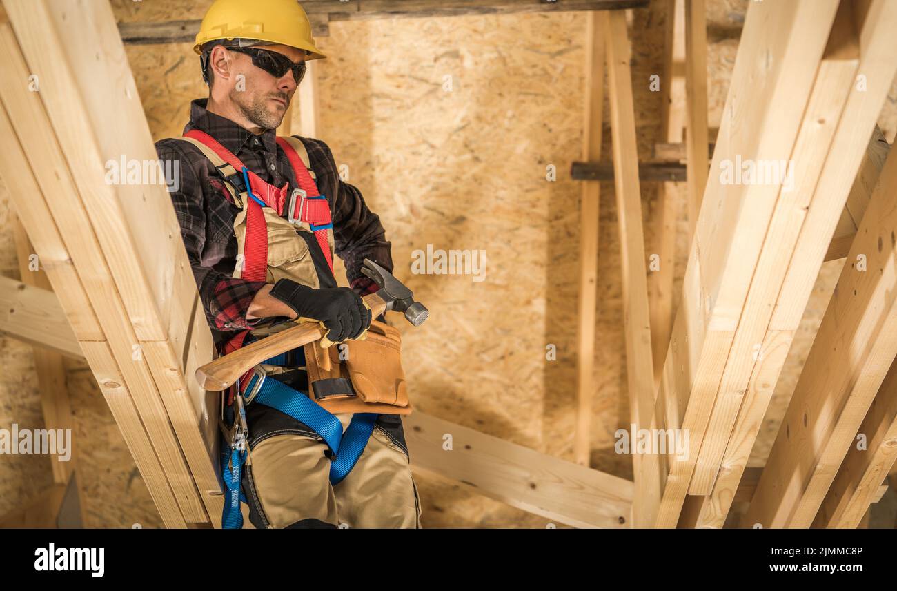Kaukasischer Baumeister mittleren Alters in gelbem Helm, Arbeitskleidung und Sicherheitsgeschirr in der Mitte des Holzwohnhauses stehen Baustelle w Stockfoto