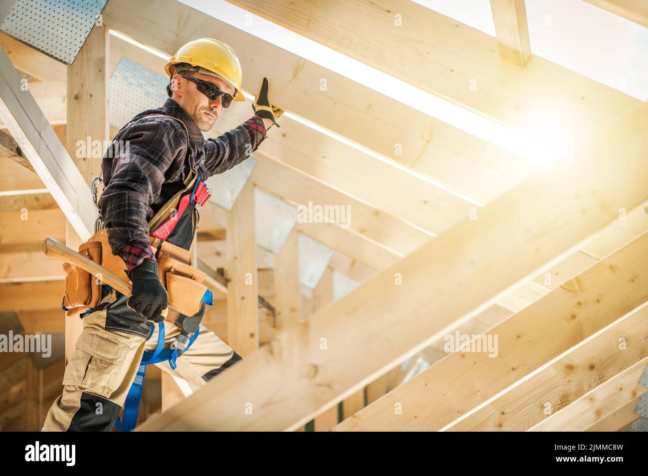 Voll ausgestatteter professioneller Bauarbeiter, der Holzbretter im Dachabschnitt-Skelett des neuen Wohnhauses sichert, indem er Nägel mit einem Ha hammend Stockfoto