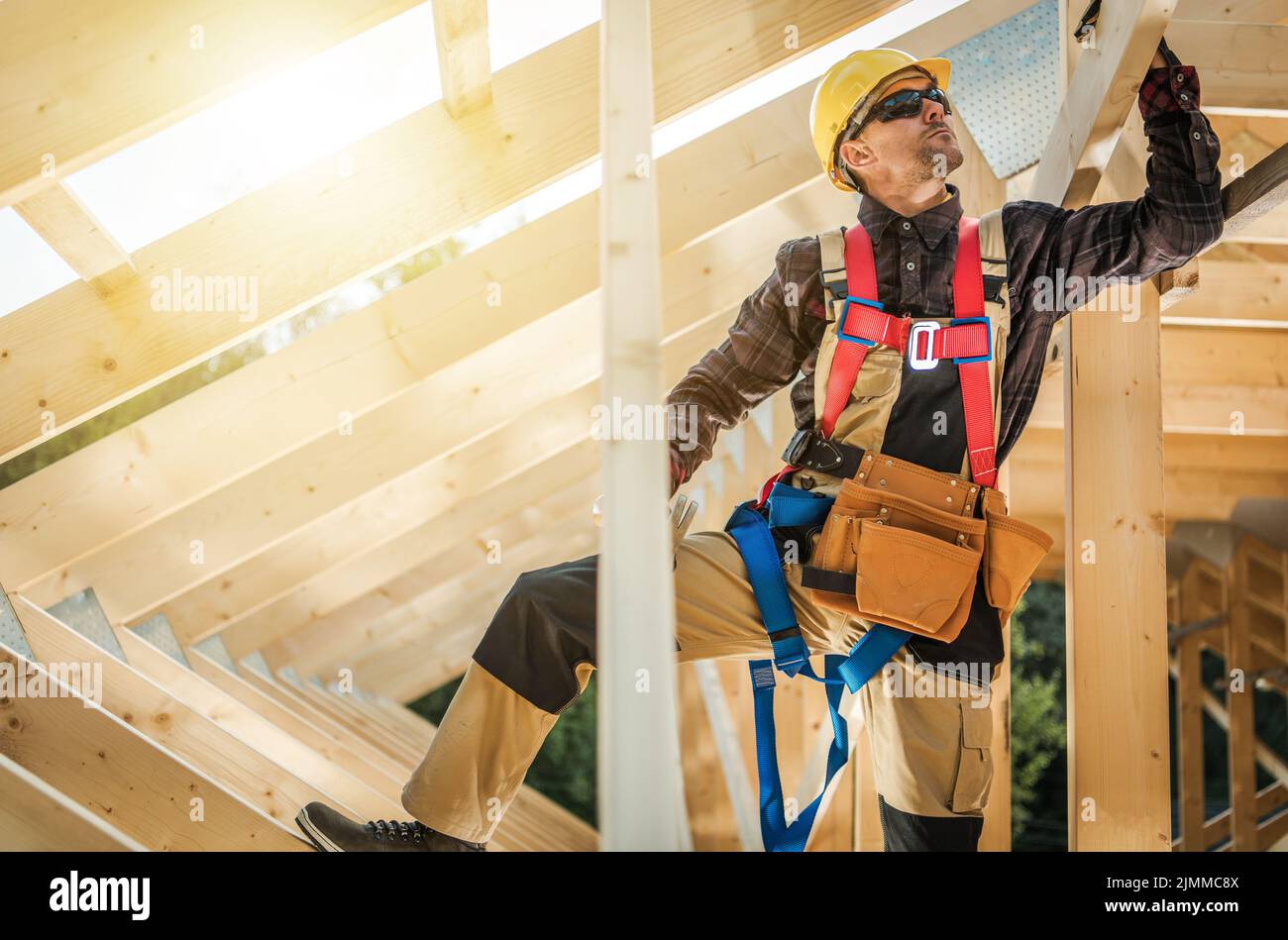 Kaukasischer Auftragnehmer, der Sicherheitskabelbaum und Schutzhelm trägt, überprüft die Festigkeit der Installation von Holzbohlen im Dachabschnitt eines Holzresims Stockfoto