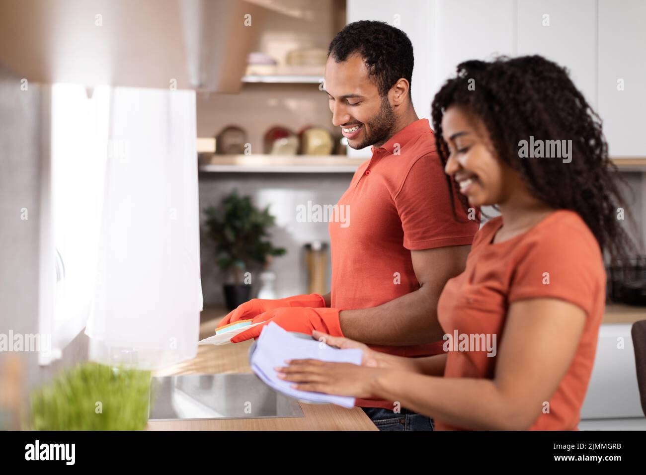 Frohes, tausendjähriges afroamerikanisches Männchen und eine Dame in den gleichen T-Shirts, die Geschirr waschen, genießen Sie die Zeit zusammen Stockfoto