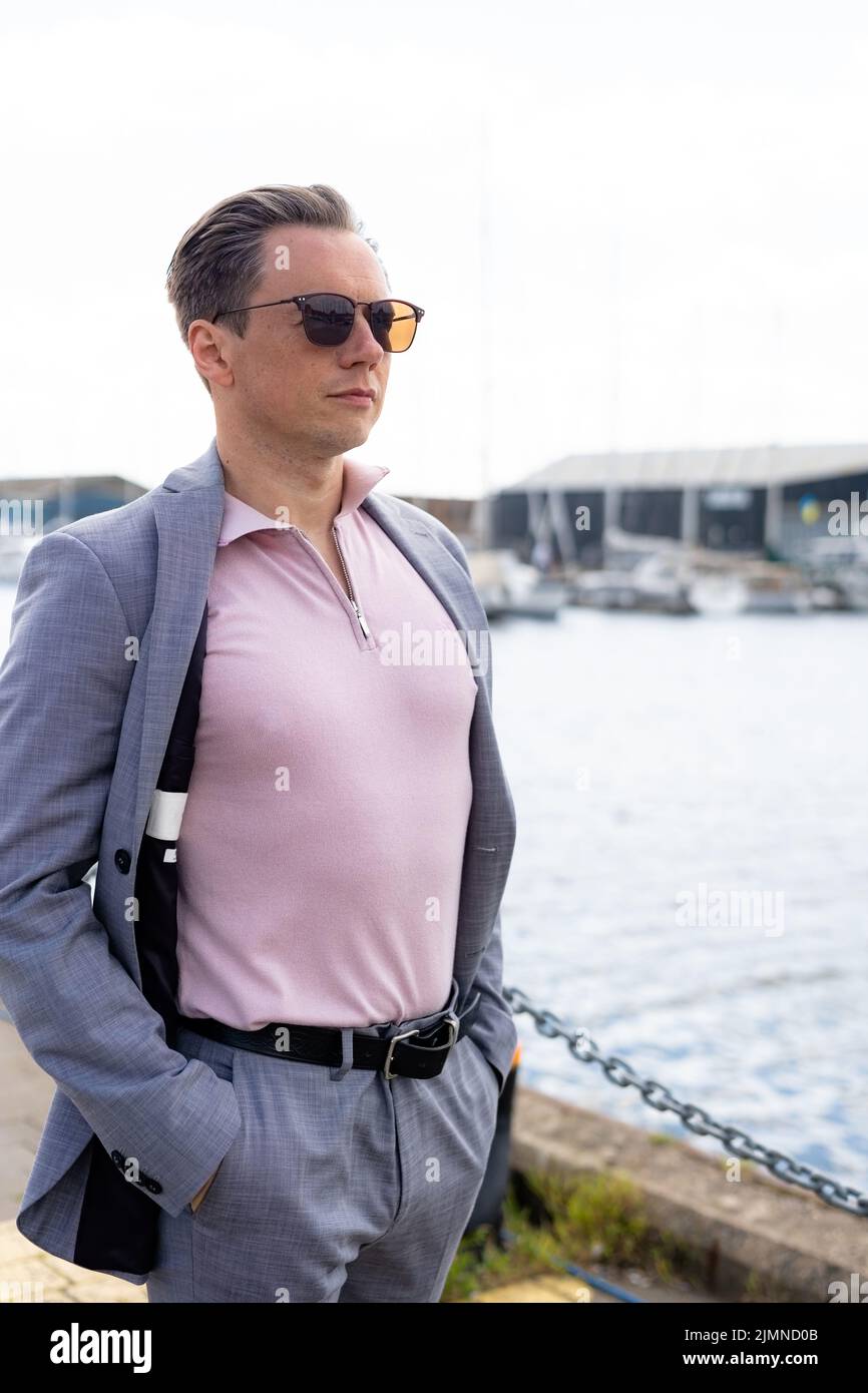 Ein hübscher Geschäftsmann in einem legeren grauen Anzug vor einem geschäftigen Yachthafen mit Luxusyachten, die vor Anker liegen Stockfoto