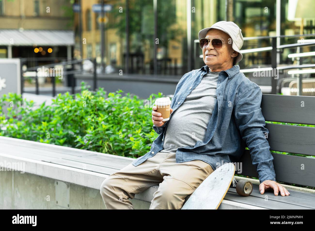 Älterer Mann mit Longboard sitzt auf der Bank und trinkt Kaffee in einem Stadtpark Stockfoto