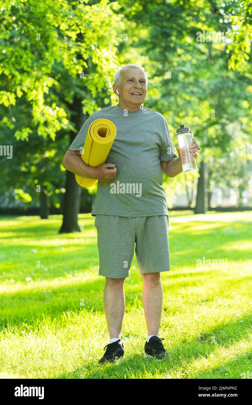 Aktiver älterer Mann mit einer Trainingsmatte und einer Flasche Wasser im grünen Stadtpark Stockfoto