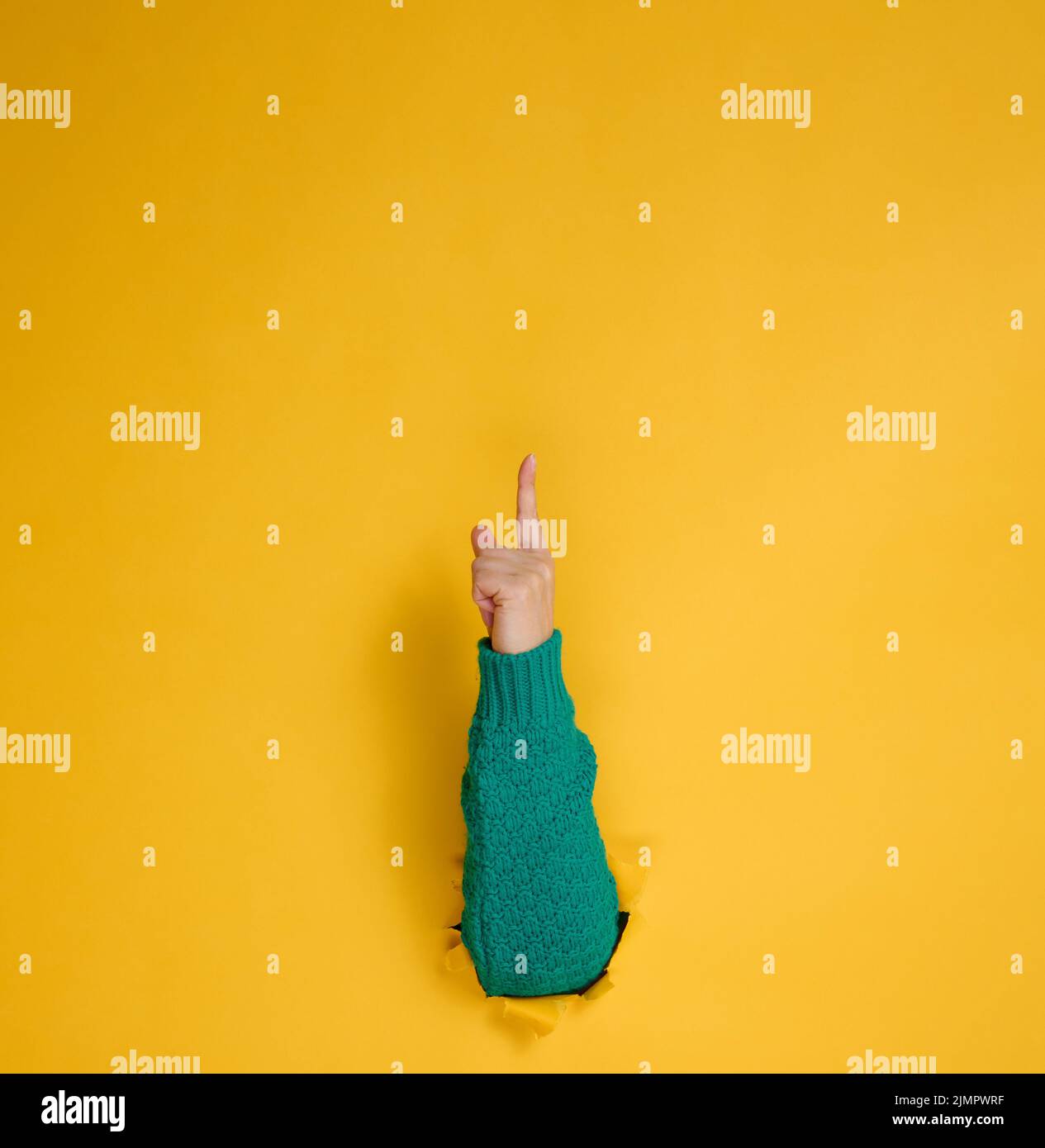 Weibliche Hand ragt aus einem zerrissenen Loch auf einem gelben Papierhintergrund, ein Teil des Körpers zeigt mit dem Zeigefinger nach oben. Kopie en Stockfoto