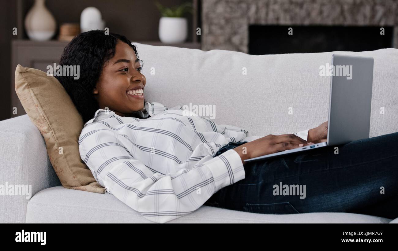 Afrikanisches emotionales Mädchen Amerikanerin, die zu Hause auf dem Sofa liegend einen Laptop ansieht, freut sich über Überraschung schockierend, dass das Wunder gute Nachrichten liest, wird bemerkt Stockfoto