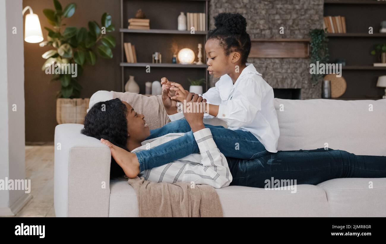Afroamerikanische liebevolle Mutter Mama, die auf dem Sofa liegt und sich zu Hause im Wohnzimmer entspannt, während sich kleine Tochter unterhielt und Spaß beim Spielen hatte Stockfoto