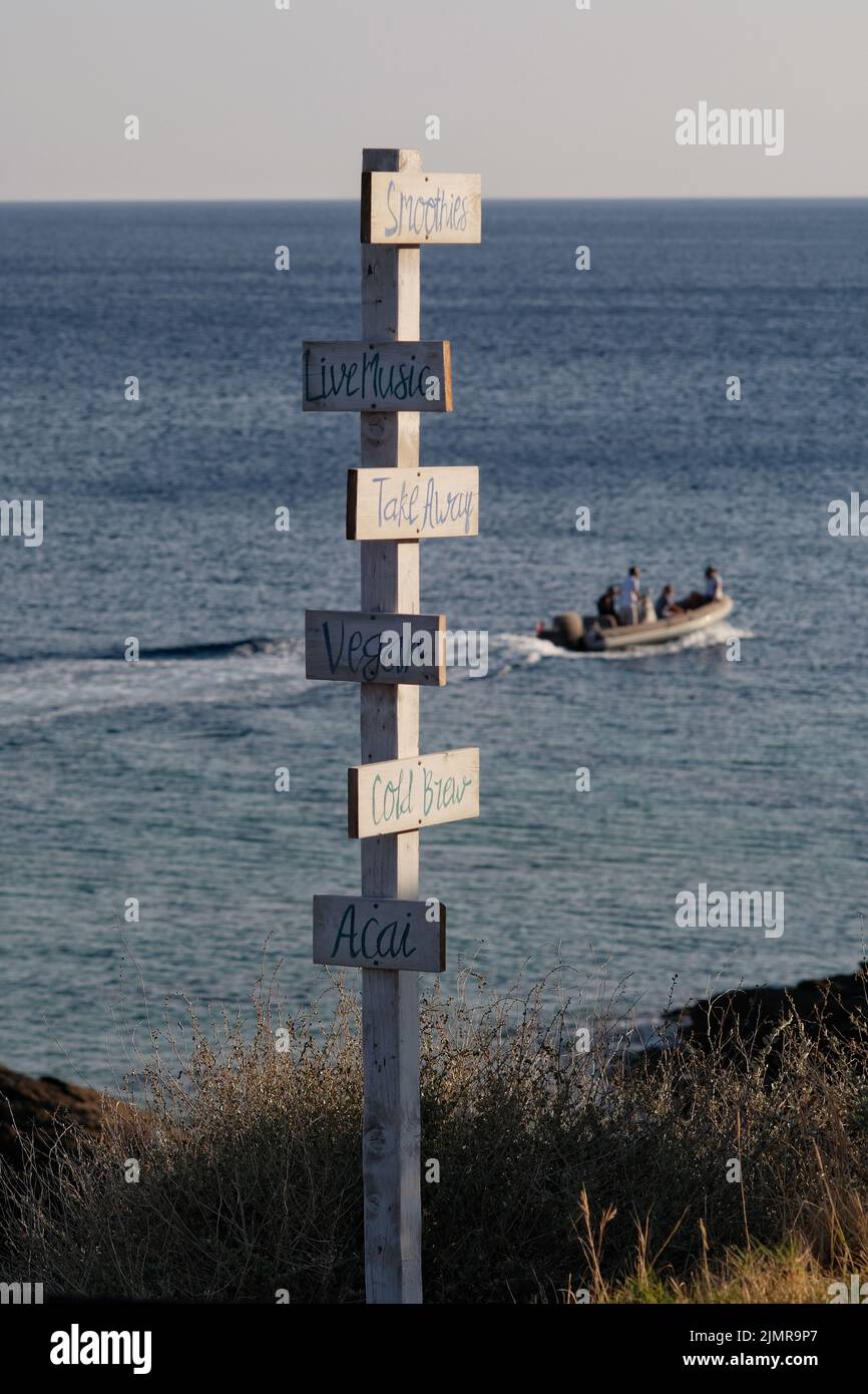 Blick auf hölzerne Schilder für Take Away, Live-Musik, acai, kaltes Gebräu, vegan, Smoothies und das Meer im Hintergrund am Strand von Mylopotas iOS Greece Stockfoto