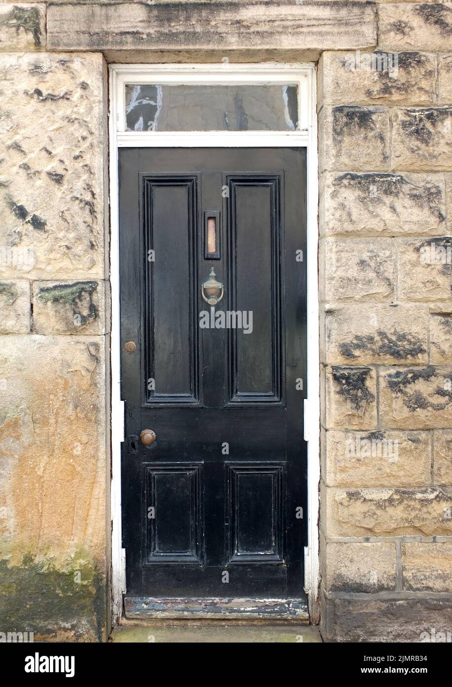 Traditionelle alte schwarz bemalte Holzhaustür mit Messingbrieffachkasten und Klopfer mit einem weißen Rahmen, umgeben von einer Steinmauer Stockfoto