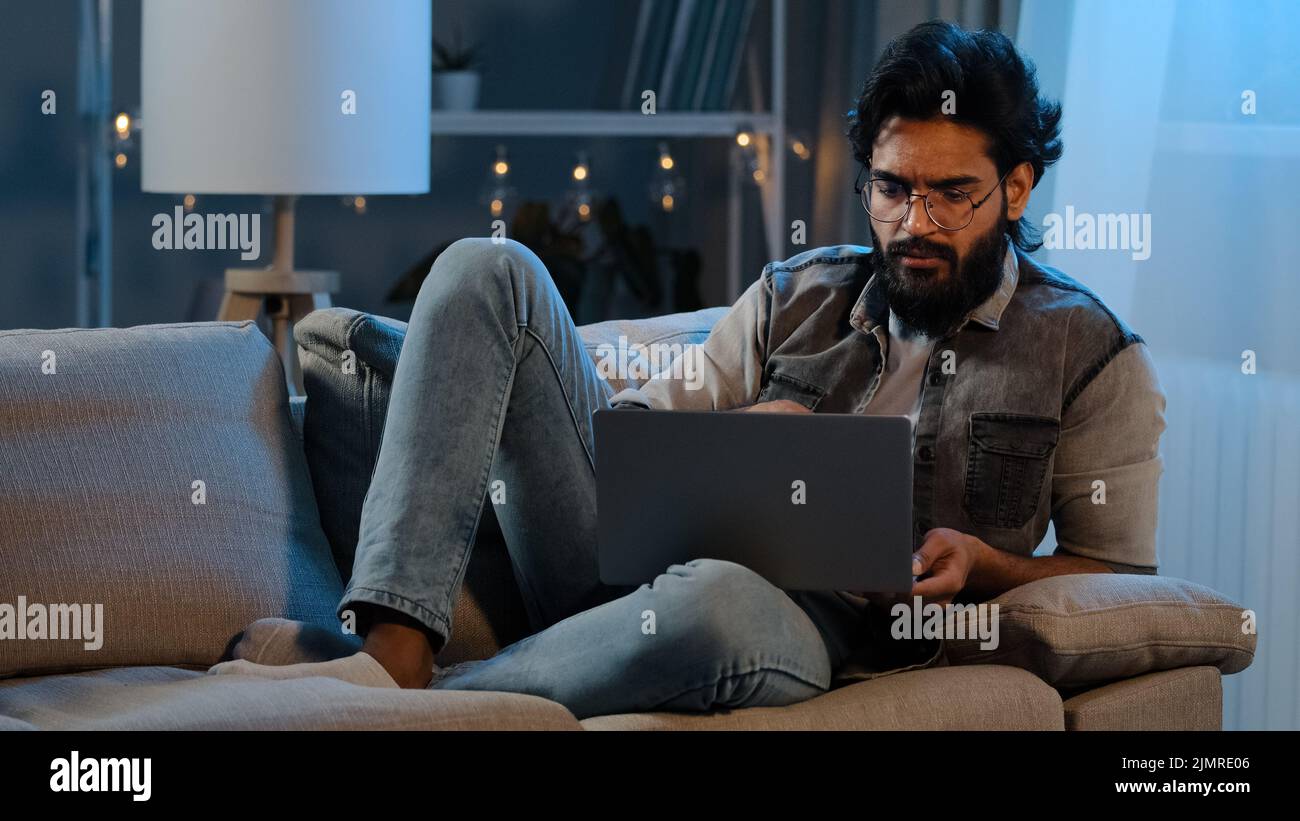 Freiberufler Typ Araber Inder Mann bärtig männlichen Benutzer trägt Brille sitzt auf dem Sofa in der Nacht spät mit Laptop Lesen Nachrichten online denken Zweifel Stockfoto