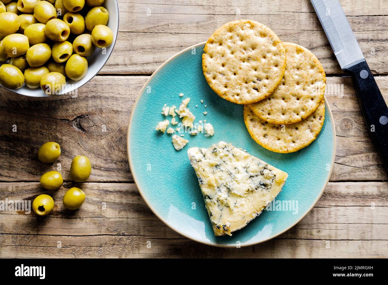 Eine Scheibe reifen Stilton-Käse auf einem Teller mit runden Weizencrackern und grünen Oliven in einer kleinen Schüssel zur Seite. Eine Portion in Wüstengröße Stockfoto
