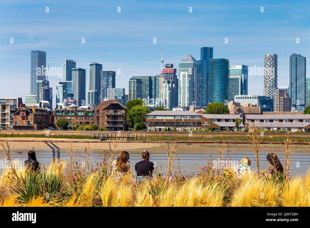 Menschen, die am Ufer des Greenwich mit Blick auf die Wolkenkratzer von Canary Wharf sitzen, London, Großbritannien Stockfoto
