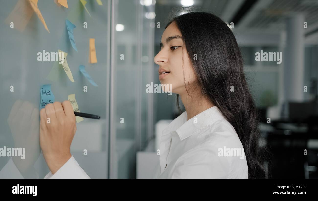 Nachdenkliche junge Geschäftsfrau arabische hispanische kreative Designerin kommen zu Notizbrett im Büro lesen bunte Aufkleber schreiben Erinnerungen auf klebrige Papiere Stockfoto