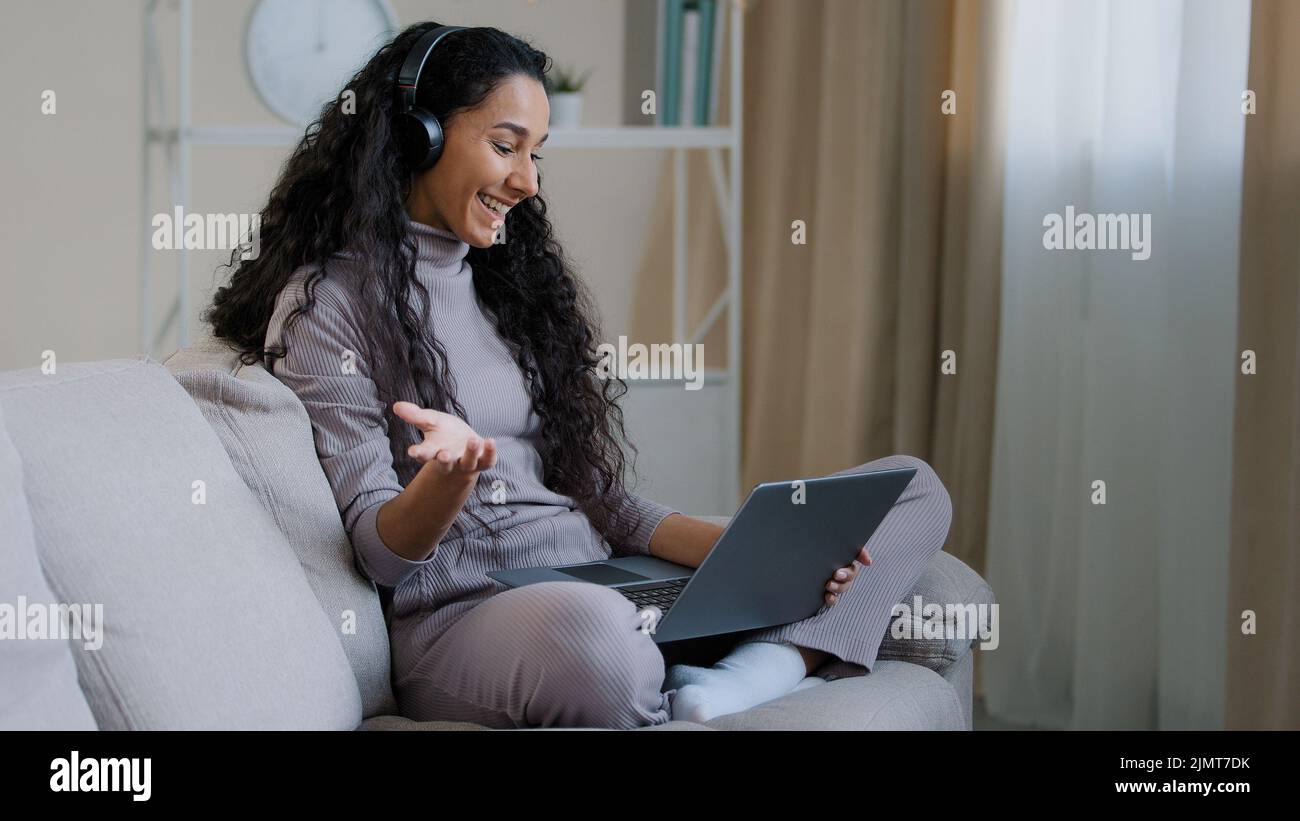 Junge hispanic arab Attraktive Freiberuflerin mit lockiger Frisur Tragen Sie ein Headset Sitzen Sie in Lotushaltung auf einem gemütlichen Sofa Halten Sie Laptop Sprechdistanz drahtlos Stockfoto