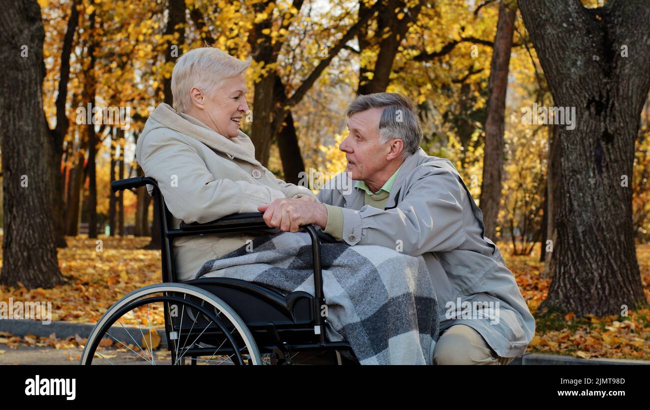 Kaukasischer älterer Mann mit weiblicher Hand fürsorgliche Frau mit Behinderung auf Rollstuhl Ehemann begleitet kranke Frau während der Rehabilitation im Herbst Park Stockfoto