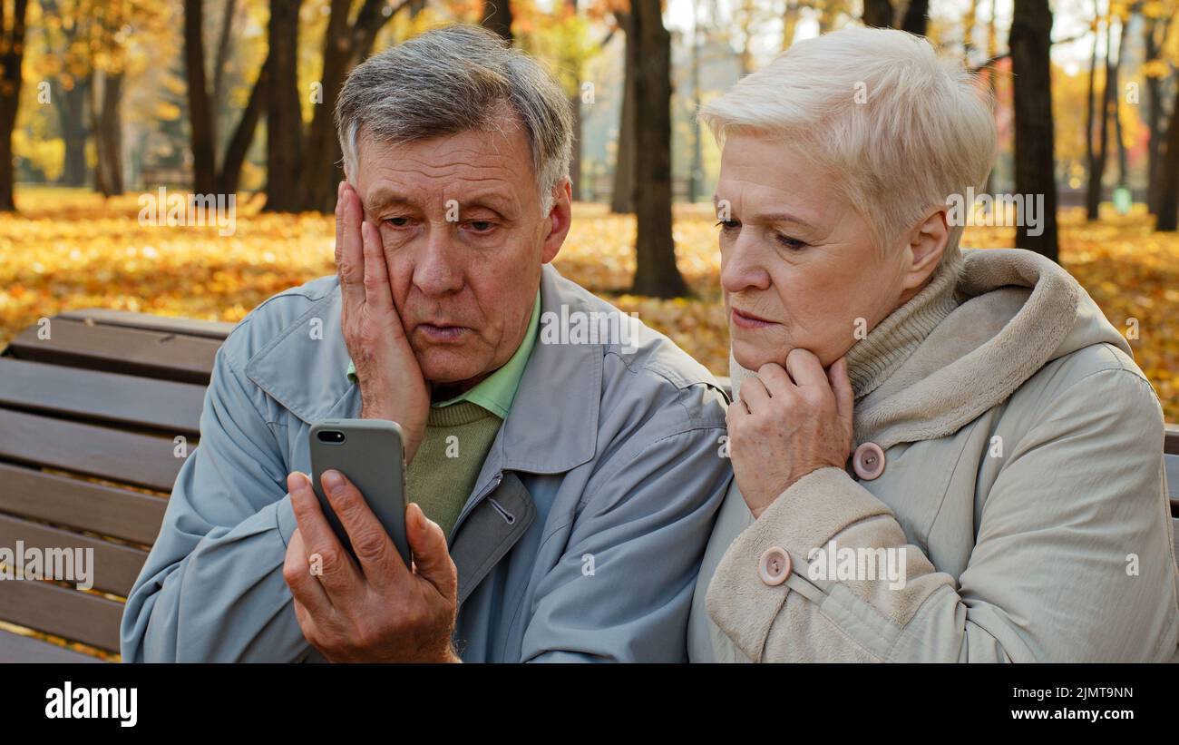 Verärgerte ältere Ehepaare sitzen im Herbstpark auf der Bank schlechte Nachrichten auf dem Smartphone lesen besorgte alte Menschen schauen sich das Bildschirmtelefon an trauriges Familiengefühl Stockfoto