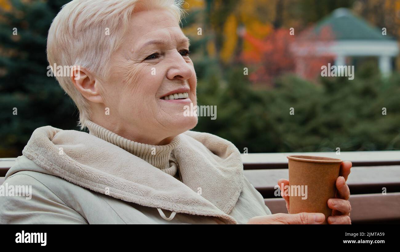 Glückliche grauhaarige traumhafte ältere Frau Großmutter trinkt Kaffee heißen Tee aus Einweg-Tasse sitzen auf Parkbank Senior gesund fröhlich Stockfoto