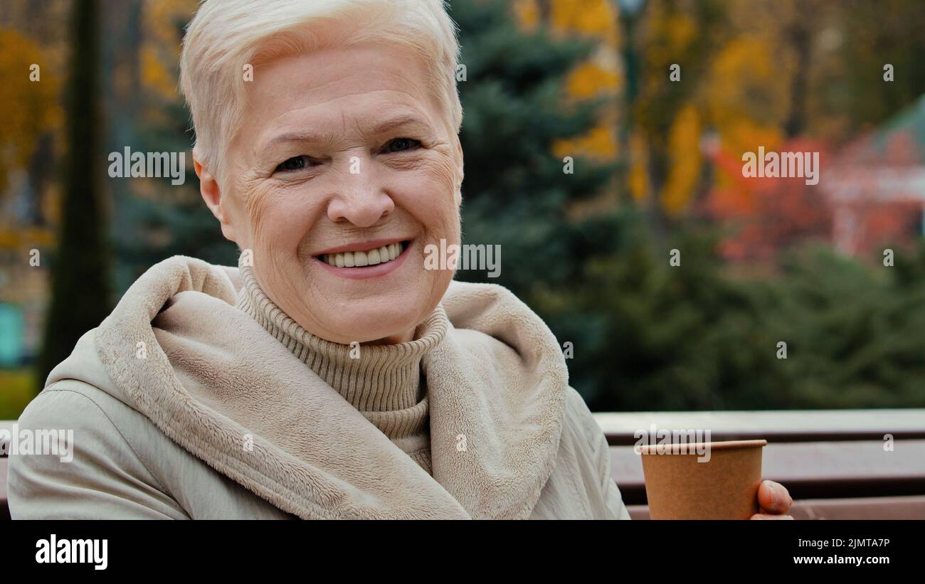 Sorglos verträumt ältere ältere Frau Oma ruht im Park trinken heißen aromatischen Kaffee aus Einweg-Tasse mittleren Alters 60s Dame sitzen auf der Bank genießen Stockfoto