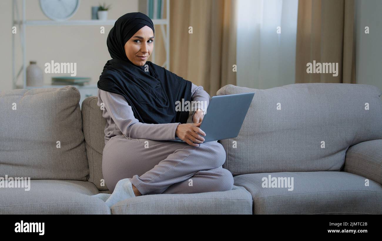 Attraktive junge muslimische Frau tragen Hijab sitzen auf gemütlichem Sofa im Wohnzimmer liebenswert energisch Millennial islamischen arabischen Mädchen hat Spaß im Wohnzimmer Stockfoto