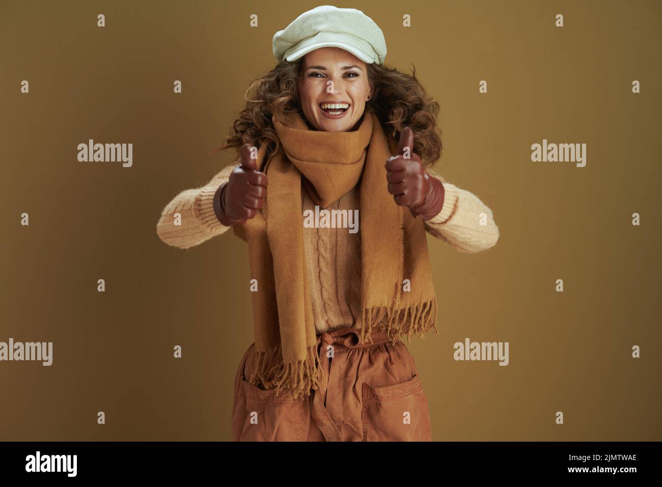 Hallo oktober. Glücklich stilvolle Hausfrau in Schal mit Lederhandschuhen zeigt Daumen nach oben auf beigem Hintergrund. Stockfoto