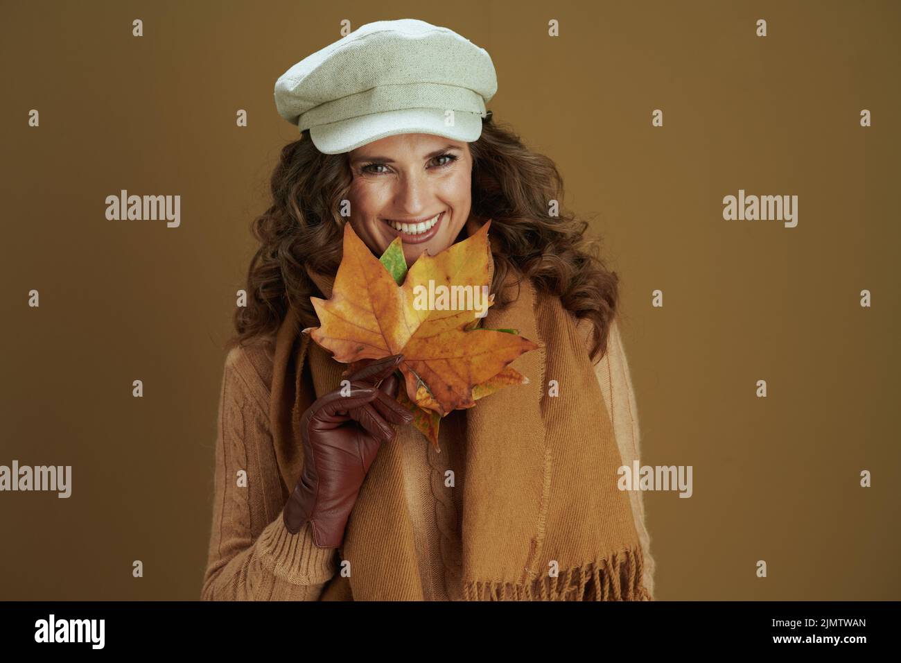 Hallo september. Porträt einer lächelnden modernen Frau in Pullover mit Lederhandschuhen und gelben Herbstahornblättern isoliert auf braunem Hintergrund. Stockfoto