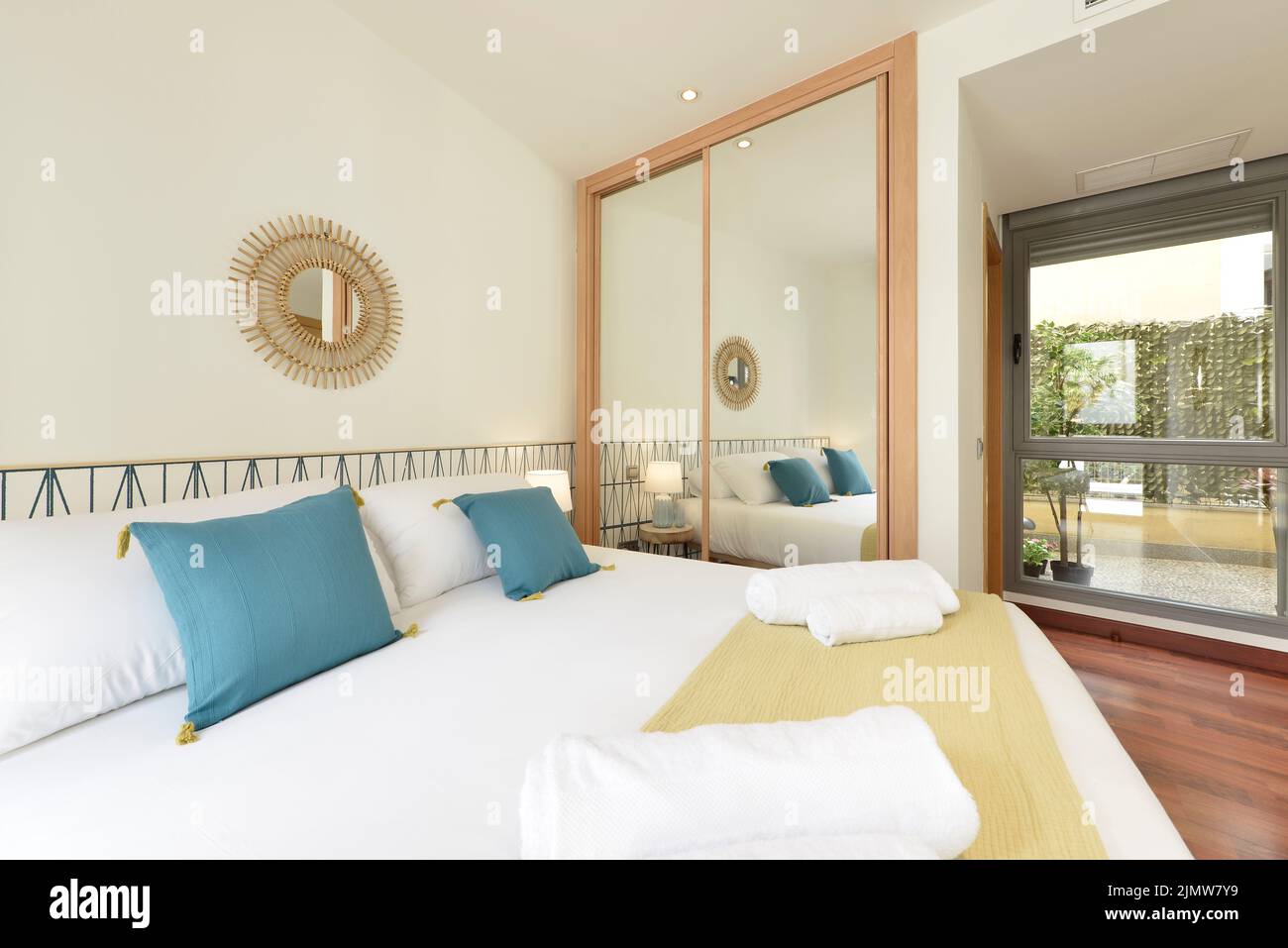 Schlafzimmer mit moderner Dekoration mit Einbauschrank mit Spiegelschiebetüren und Holzkanten, dekorativen Spiegeln, blauen Kissen und einem Fenster Stockfoto